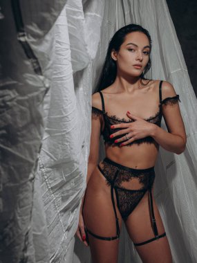 Dantelli siyah iç çamaşırlarıyla poz veren genç seksi esmer kadın ve gri arka planda karanlık stüdyoda pantolon askısı. BDSM tarzı. Yakın plan..
