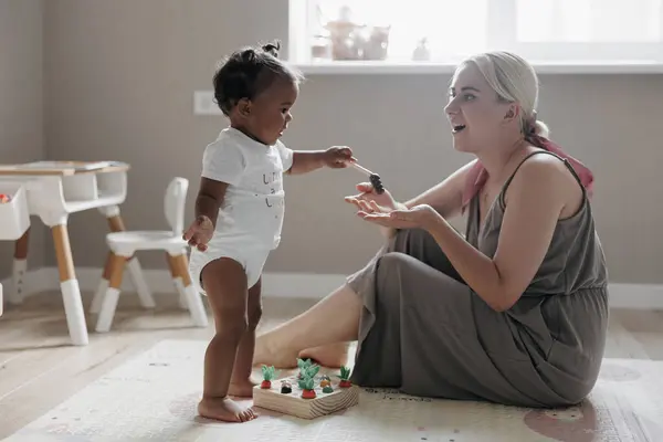 Happy caucasian mother sits on floor in nursery and plays with her cute mixed race toddler daughter. Concept of interracial family.