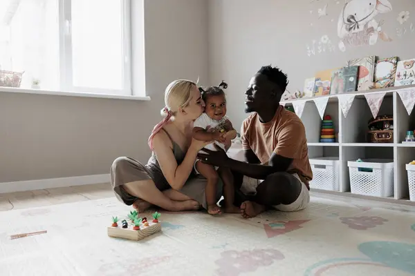 Happy multiracial parents playing with their toddler daughter in nursery. Parents kissing and embracing their daughter. Concept of interracial family.