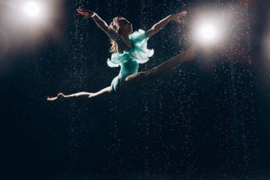 Yeşil elbiseli balerin kız, düşen su damlalarına ve arka plandaki spot ışıklarına karşı jimnastik hareketler yapıyor. Zarif balerin. Bale sanatı, zarafet, esneklik.