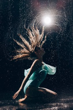 Yeşil elbiseli genç balerin kız suya düşen suyun altında dans ediyor ve siyah arka planda ıslak saçlarını spot ışıklarına doğru sallıyor. Zarif balerin. Bale sanatı, zarafet, esneklik.