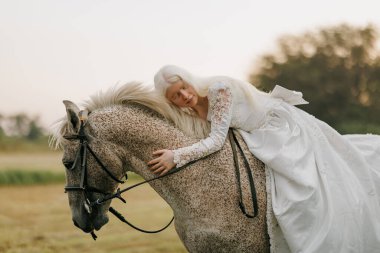 Uzun beyaz elbiseli genç albino kadın ata biner ve gri ata sarılır gökyüzüne ve çayır arka planına.