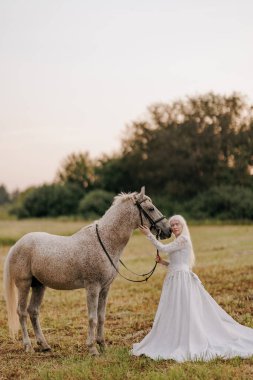 Uzun beyaz elbiseli genç albino kadın gri atın yanında duruyor ve gün batımının arka planında çayırda sarılıyor..