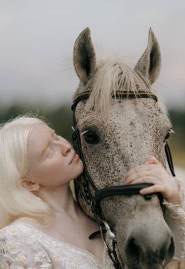 Beyaz elbiseli genç albino kadının portresi gri atın yanında duruyor ve çayırda sarılıyor..