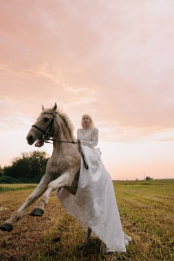 Uzun beyaz elbiseli genç albino kadın günbatımı gökyüzünün arka planında arka planda gri at üzerinde oturuyor..