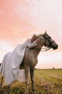 Uzun beyaz elbiseli genç albino kadın ata biniyor ve gün batımının arka planında gri ata sarılıyor..