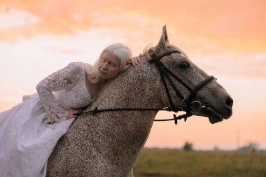Genç albino kadının yakından görüntüsü beyaz elbiseli gri atın sırtında ata biner ve günbatımı gökyüzünün arka planında sarılır..