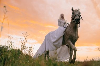 Uzun beyaz elbiseli genç albino kadın gün batımının arka planında gri ata biniyor..