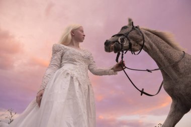 Beyaz elbiseli genç albino kadının portresi gün batımının arka planında çayırda gri atı dizginliyor. Alt görünüm.