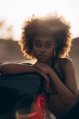 Gün batımında kumlu kanyonun arka planında siyah sutyenli açık kamyonet yatağında oturan Afrikalı güzel bir kadın. Portre.
