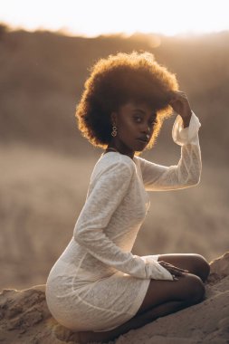 Kumlu Kanyon 'da beyaz elbiseli güzel bir Afrikalı kadın gün batımının arka planında oturuyor. Arka ışıklandırma. Yakın çekim..