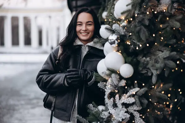 Ceketli ve eldivenli genç bir kadın Noel ağacının yanında duruyor. Parlayan çelenkler ve toplarla süslenmiş..