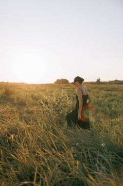 Siyah elbiseli ve güneş gözlüklü genç bir kadın gün batımında otların arasında çimenlerde yürüyor. Arkaplan.