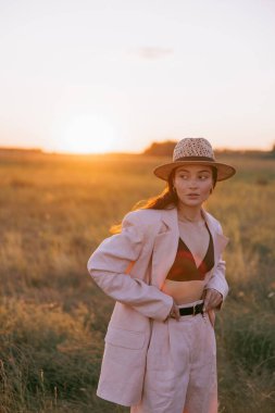 Siyah sütyenli, ceketli ve hasır şapkalı genç bir kadın gün batımında otların arasında çayırda yürüyor. Arka ışık. Yakın plan..