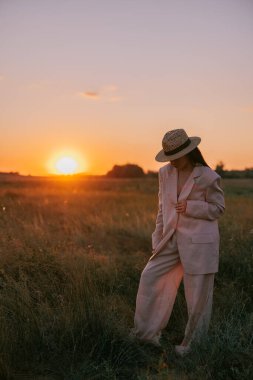 Siyah sutyen, ceket, pantolon ve hasır şapkalı genç bir kadın gün batımında otların arasında çayırda yürüyor. Arkaplan.