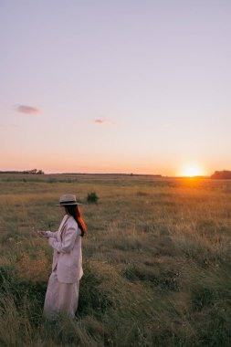 Siyah sutyen, ceket, pantolon ve hasır şapkalı genç bir kadın gün batımında otların arasında çayırda yürüyor. Arkaplan.