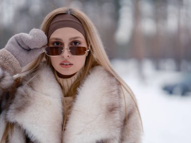 Karlı kış ormanlarında çam ağaçlarının arkasında yürürken kürk ceketli ve güneş gözlüklü güzel bir kadının portresi..