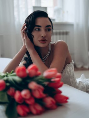 İnci kolyeli seksi esmer kadın portresi ve yatağın yanında oturan erotik saten tulum ve kırmızı lale buketi. Erotik kadın iç çamaşırı. Düğün için erotik iç çamaşırı..