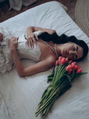 İnci kolyeli seksi esmer bir kadın ve erotik bir vücut giysisi. Yatakta uzanmış, gözleri kapalı bir şekilde dinleniyor. Bir sonraki kırmızı lale buketi. Erotik kadın iç çamaşırı. Düğün için erotik gelinlik. Üst görünüm.