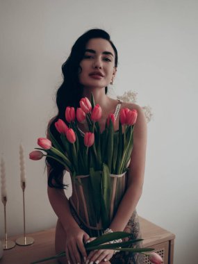 Bacağında dövmesi olan seksi esmer kadın inci kolye ve bilezik takmış kırmızı lalelerle poz veriyor. Yakın plan..