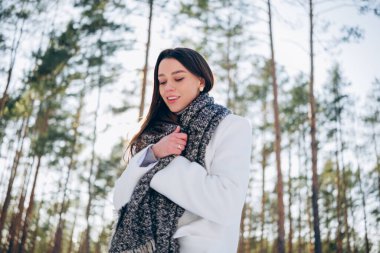 Kışın yürürken çam ağaçlarının arasında beyaz önlüklü ve eşarplı esmer kadın portresi.