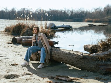 Trençkotlu genç kadın gölün kıyısındaki kütükte oturuyor. Sakin suları gökyüzünü, bulutları ve ilkbaharın başlarında çıplak ağaçlarla çevrili..