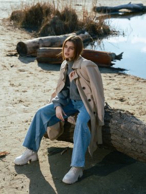 Trençkotlu genç bir kadın gölün kıyısındaki kütükte oturur. Sakin suları, baharın başlarında gökyüzünü ve bulutları yansıtır. Yakın plan..