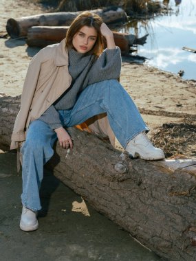 Trençkotlu genç bir kadın gölün kıyısındaki kütükte oturur. Sakin suları, baharın başlarında gökyüzünü ve bulutları yansıtır. Yakın plan..