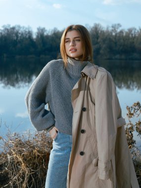Trençkotlu genç kadın göl kenarında duruyor. Sakin suları gökyüzünü, bulutları ve ilkbaharın başlarında çıplak ağaçlarla çevrili. Yakın plan..