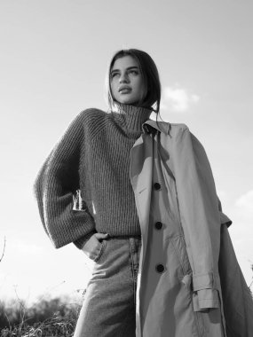 Trençkotlu genç kadın gökyüzü ve bulutların arkasında duruyor. Yakından ve alttan. Siyah ve beyaz resim.