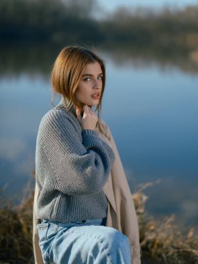 Süveter giymiş genç düşünceli bir kadın, sakin sularıyla gölün kenarında durur ve baharın başlarında gökyüzünü ve bulutları yansıtır. Yakın plan..