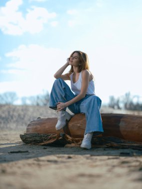 Beyaz atletli ve kot pantolonlu genç esmer kadın kumsalda, bulutlu arka planda oturuyordu..