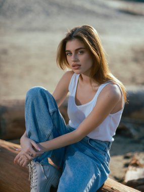 Beyaz atletli esmer kadın portresi ve kumsalda oturan kot pantolon..