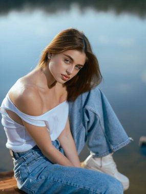 Beyaz kolsuz tişörtlü genç bir kadın portresi ve gölün kenarında oturan ve rahatlatan bir kot pantolon. Gökyüzünü bulutlarla yansıtan sakin sular..
