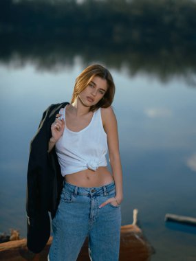 Beyaz kolsuz tişörtlü, kot pantolonlu, kumsalda, odunların yanında, göl kıyısında, gölü, gökyüzünü bulutları ve ağaçları yansıtan genç bir kadın. Yakın plan..