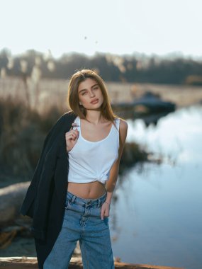 Beyaz kolsuz tişörtlü, kot pantolonlu ve göl kıyısında duran sakin suları gökyüzünü ve bulutları yansıtan genç bir kadın. Yakın plan..