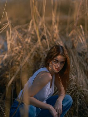 Beyaz atletli genç esmer kadın ve kuru kamışların arasında oturan ve rahatlatan kot pantolon. Yakın plan..