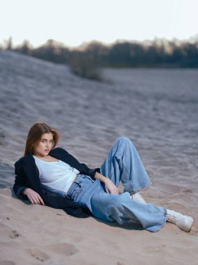 Beyaz atletli genç esmer kadın, kot pantolon ve ceket kumsalda uzanmış, kumların ve gökyüzünün arkasında..