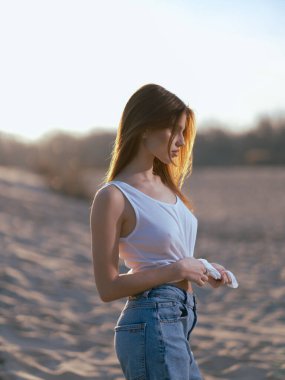 Beyaz atletli ve kot pantolonlu genç esmer kadın gün batımında kumların ve gökyüzünün arka planında duruyor. Yakın plan. Yan görünüm.