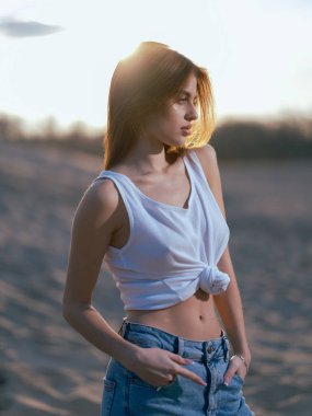 Beyaz atletli ve kot pantolonlu genç esmer kadın gün batımında kumların ve gökyüzünün arka planında duruyor. Yakın plan..