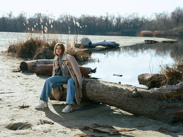 Trençkotlu genç kadın gölün kıyısındaki kütükte oturuyor. Sakin suları gökyüzünü, bulutları ve ilkbaharın başlarında çıplak ağaçlarla çevrili..