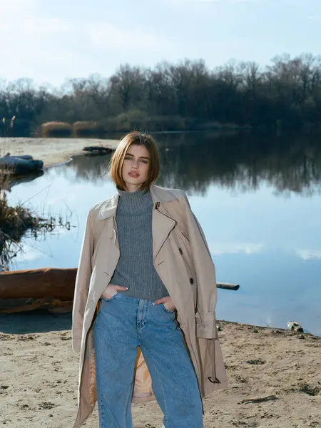 Trençkotlu genç bir kadın gölün kıyısında duruyor. Durgun suları gökyüzünü, bulutları ve ilkbaharın başlarında çıplak ağaçlarla çevrili..
