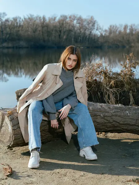 Trençkotlu genç kadın gölün kıyısındaki kütükte oturuyor. Sakin suları gökyüzünü, bulutları ve ilkbaharın başlarında çıplak ağaçlarla çevrili..