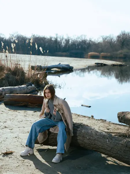 Trençkotlu genç kadın gölün kıyısındaki kütükte oturuyor. Sakin suları gökyüzünü, bulutları ve ilkbaharın başlarında çıplak ağaçlarla çevrili..