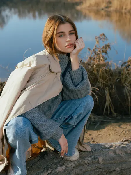 Trençkotlu genç bir kadın gölün kıyısındaki kütükte oturur. Sakin suları, baharın başlarında gökyüzünü ve bulutları yansıtır. Yakın plan..