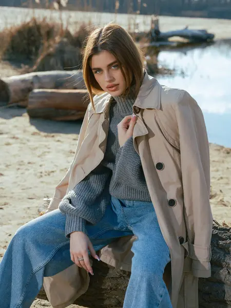 Trençkotlu genç bir kadın gölün kıyısındaki kütükte oturur. Sakin suları, baharın başlarında gökyüzünü ve bulutları yansıtır. Yakın plan..