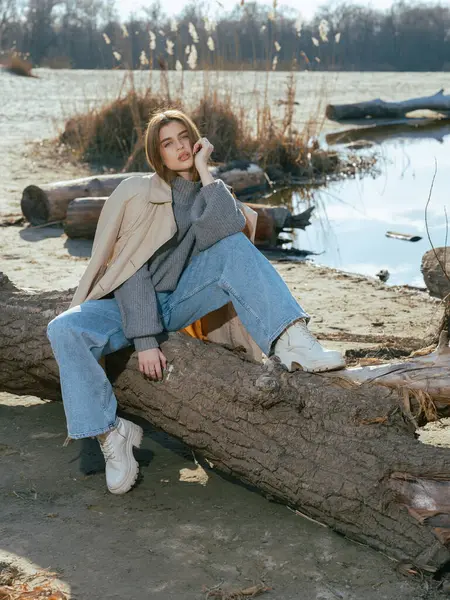 Trençkotlu genç kadın gölün kıyısındaki kütükte oturur. Sakin suları gökyüzünü ve baharın başlarındaki bulutları yansıtır..