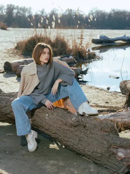 Trençkotlu genç kadın gölün kıyısındaki kütükte oturur. Sakin suları gökyüzünü ve baharın başlarındaki bulutları yansıtır..