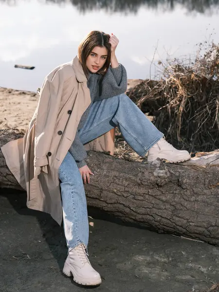 Trençkotlu genç bir kadın gölün kıyısındaki kütükte oturur. Sakin suları, baharın başlarında gökyüzünü ve bulutları yansıtır. Yakın plan..