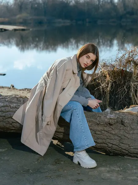Trençkotlu genç bir kadın gölün kıyısındaki kütükte oturur. Sakin suları, baharın başlarında gökyüzünü ve bulutları yansıtır. Yakın plan..